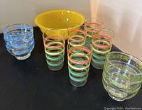 Five striped glasses with horizontal pastel colored bands, three stacked blue floral pattern bowls, and large yellow Pyrex serving bowl on table