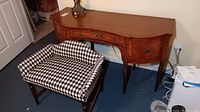 Antique vanity with painted floral detail and three drawers, showing some nicks, along with an upholstered bench in black and white checkered fabric showing damage to upholstery.