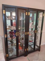 Front view of two adjacent black Asian style lighted curio cabinets filled with display items, showing brass hardware and glass shelving.