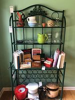Front view of the black metal bakers rack showing all four shelves filled with various glassware, kitchen items and cookbooks to illustrate rack capacity. Also visible is the decorative top panel and integrated wine holder underneath the lowest shelf.