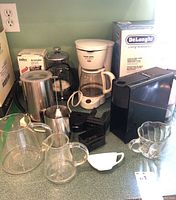 Wide view showing multiple coffee related items including a Nespresso machine, drip coffee maker, French press, glass pitchers, frother, and black plastic accessories on a countertop.