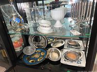 Photo showing blue floral crystal decanter, glass items labeled 'Matzo', milk glass compote, brass menorah, and pewter/judaica serving pieces on two glass shelves.