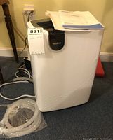 Photo showing the front view of the dehumidifier with a tag and coiled hose beside it on carpeted floor.