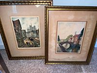 Two framed colored engraving prints showing European scenes with gilt frames and beige mats. Frames and mats show visible water damage and age.