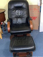 Front view of the black leather reclining glider chair with its wooden armrests and base, showing the cushioned back and seat with some wear.