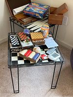 Two black metal game tables with grid-style tops and a variety of board games and accessories arranged on top showing signs of light use and some vintage condition.