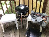 View of the portable charcoal grill between two white plastic side tables with grilling tools laid out on one table and a black grill grate on the wooden deck below.