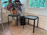 Two black metal patio side tables, a faux plant in a terracotta pot, and a black metal urn on a tiled floor near windows.