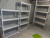 Three taller plastic shelving units each with four slatted shelves, beige/off-white color, placed against a concrete wall. Condition shows dirt and some cracking.