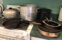 Photo showing a collection of various pots and pans including cast iron skillet, large aluminum pots, black enamel pot, copper pan and other cookware placed on stove and counter.