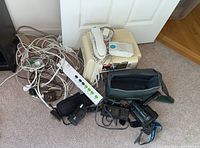 Photo showing multiple power cords, power strips, a cream-colored landline phone, camera and camcorder carrying cases on a wooden floor.