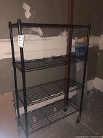 Front view of black metal rolling shelf unit, showing four wire mesh shelves and caster wheels.