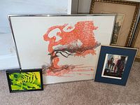 Photo shows three framed art pieces: a large silkscreen print with a red-orange abstract tree, a framed signed photo with red and black abstract shapes under blue matting, and a small green and yellow foliage print.