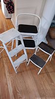 Set of 3 step stools arranged on wood floor: two folding white stools with black steps and one fixed black step stool
