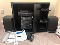 Full view of stereo system, VHS player and speakers within wooden media cabinet, also shows user manuals and two remote controls on carpet in front.