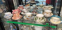 Full view of assorted china tea cups and saucers arranged on glass shelf