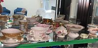 Wide view of multiple vintage tea cups with saucers and a china bowl on a glass shelf, showing variety and condition including chips and repairs.