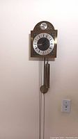 Frontal view of the full clock mounted on a wall showing the pendulum weight and chain system.