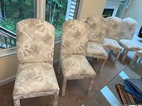 Photo showing six upholstered dining chairs arranged in a row by a window. Chairs have a floral design fabric upholstery in beige and off-white tones.