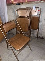Four vintage Futura folding chairs with bronze metal frames, wood grain finish seats and backrests, displayed folded and one open in basement setting.