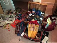 Wide shot of all knitting supplies spread out including basket of yarn, suitcase containing knitting needles, knitting patterns on floor.