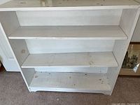 Front view of white painted wooden bookcase showing three adjustable shelves, some wear and dirt marks visible.