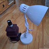 Photo of two table lamps on wood floor: white flexible-neck lamp and dark square ceramic base lamp, no shades.