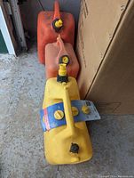 Three gas cans arranged in a line from front to back: yellow (front), orange (middle), red (back). The yellow can is new with label attached, the other two are used with wear marks.