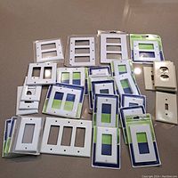 Assortment of various electrical cover plates spread out on a surface, including single, double, and triple switch covers, outlet covers, some still in original blue and green packaging, all in white color.