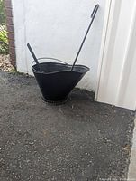 Black coal scuttle with poker and shovel placed inside on an outdoor surface.
