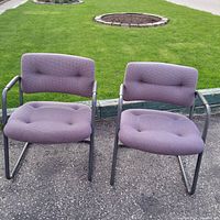 Photo of two matching purple fabric office chairs with black metal cantilever bases placed outdoors on asphalt with grass in the background.