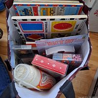 Photo of top-down view into reusable shopping bag showing children's educational board books with shapes, a ball of white yarn, plastic craft beads, and a pink box with white snowflake design.