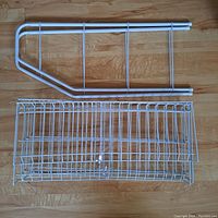 Top-down view of white metal shoe rack parts laid out on wooden floor, showing tubular frame and wire mesh basket structure.