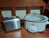 Photo showing a stainless steel two-slice toaster and a white Crock-Pot slow cooker with glass lid, placed on a wooden surface.