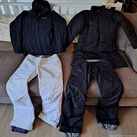 Two black coats (one Reitman 3/4 length and one Columbia 2-in-1) and two pairs of snowpants (white Columbia and black Burton) displayed on a brown couch.