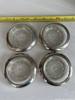 Top view of four round sterling silver coasters with clear glass inserts showing sunburst pattern and a tape measure for scale (approx 4 inch diameter each).
