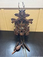 Front view of the cuckoo clock showing carved stag head with antlers above clock face, crossed rifles, leaves, and pine cone weights on chains in front.
