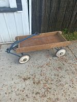 Side view of antique wooden wagon showing wooden bed, metal frame, handle, and tires on concrete surface near a white door and wooden fence.