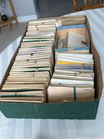 Photo showing a cardboard box filled with many envelopes containing used postage stamps organized in rows.