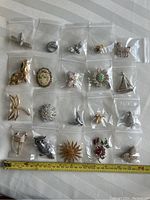 Full view of 21 individual brooches in separate plastic bags laid on white surface with measuring tape showing scale.