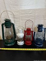 Four barn lanterns in green, white, red, and blue arranged side-by-side on a black surface with a tape measure in front showing approximately 15 inches tall for the tallest lantern.
