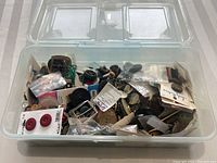Clear plastic box filled with assorted buttons and sewing materials, some brand new on cards, others loose showing signs of wear.