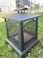 Front and top angled view of the square outdoor metal fire pit sitting on grass, showing mesh sides and chimney cap.