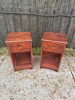 Pair of vintage wooden night stands shown side by side outdoors on concrete with bamboo fence background. Both stands have single drawer and open shelf.