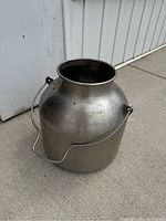 Side view of the round stainless steel milk pail with wire handle, showing surface wear and patina.