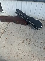 Two acoustic guitar hard shell cases, black and brown, placed side by side outside on a concrete surface showing overall worn exterior condition consistent with age and use.