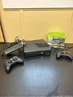 Xbox 360 console, power cable, two wireless controllers, and stack of game cases on a black surface.