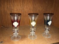 Three small glass cups on a flat surface with wood grain background. Each glass has a colored stripe and a white flower design in front.