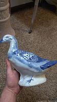 Side view of blue and white ceramic duck figurine held in hand, showing detailed feather pattern and blue painted feet and beak.