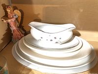 Photo of white ceramic gravy boat with subtle floral design, next to stacked white oval plates on a shelf with a small figurine.